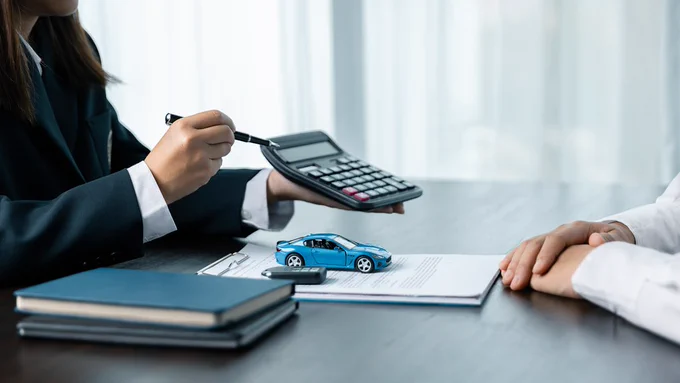 This is an image of two people talking, one is holding a calculator to the other. There is a miniature car on the table. 