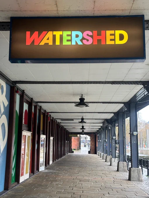 A photo of the covered walkway outside watershed with a new sign - a black light box with a rainbow coloured watershed logo 