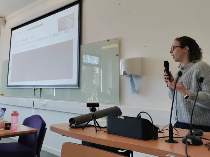 Rebecca (curly shoulder length hair, glasses, grey woolly jumper), presents on her work whilst holding a microphone and gesturing with her free hand. A powerpoint slide next to her has the title of her talk 'Legal Storytelling: Dismantling the masterplot of domestic abuse'.
