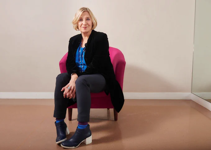 The late Victoria Wood is sitting on a comfortable chair. She is looking directly at the camera and smiling. 