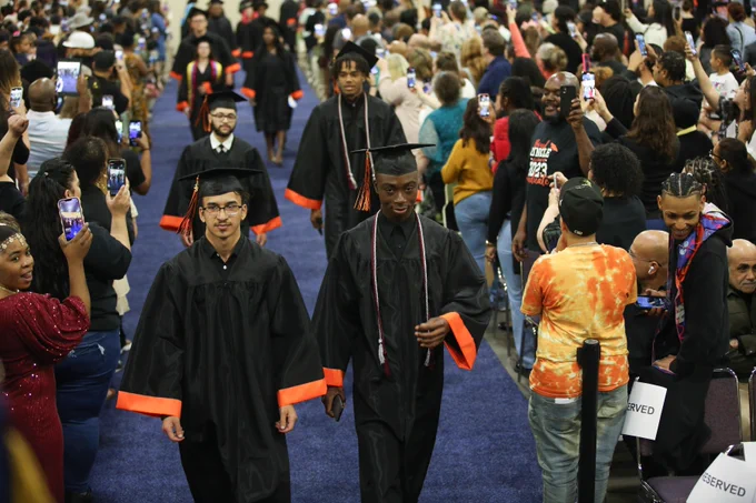 Two men lead a line of students walking down the aisle at high school graduation. 