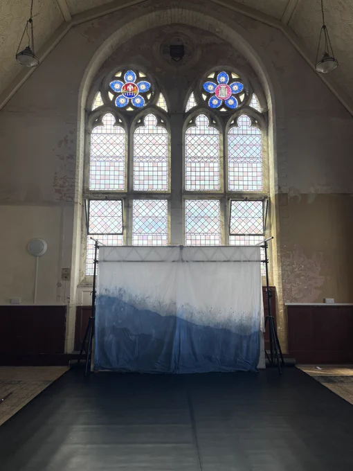 A dancefloor is set up in an old hall. There is a truss with a cotton backdrop hung over it. The backdrop is dyed blue at the bottom. Behind the truss is a large stained glass window, reaching high into the ceiling