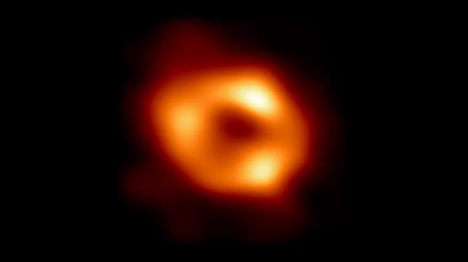 This image shows the black hole at the center of the Milky Way, Sagittarius A*. It appears as a blurry glowing orange circle with a dark hole in the middle.