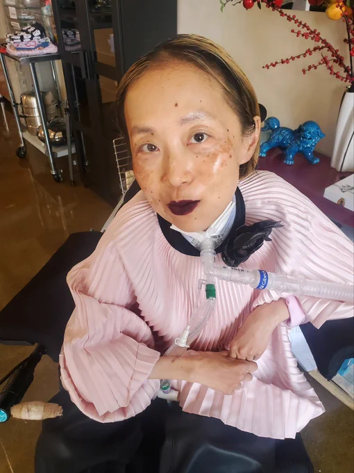 Asian American woman in a wheelchair with a tracheostomy at her neck with  a tube connected to a ventilator. She is wearing a pink flowy silky top with pleats and faux black leather pants. At the top is a black collar with a large black flower on the side. She’s giving a mysterious smile. Her lip color is dark and vampy, by Makeup Forever in never ending plum