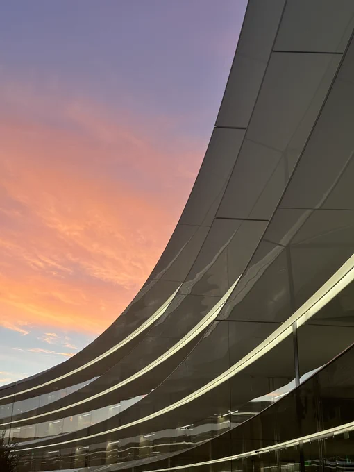 A photo of the ring building at Apple park during sunset. Architectural features of the building reflect Apple Park. The sky above is a gradient from blue to purple and the sun illuminates the clouds in a bright orange.