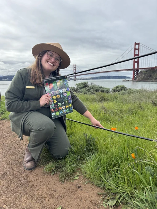 A smiling NPS Park Ranger poses for a photo with some CA Wildflowers. The Golden Gate Bridge can be seen in the background