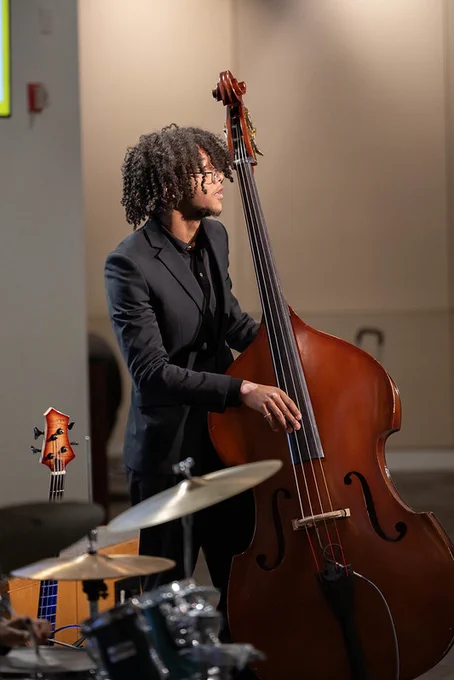 A student wearing a suit is playing a string instrument; visible in the foreground is a drum set.
