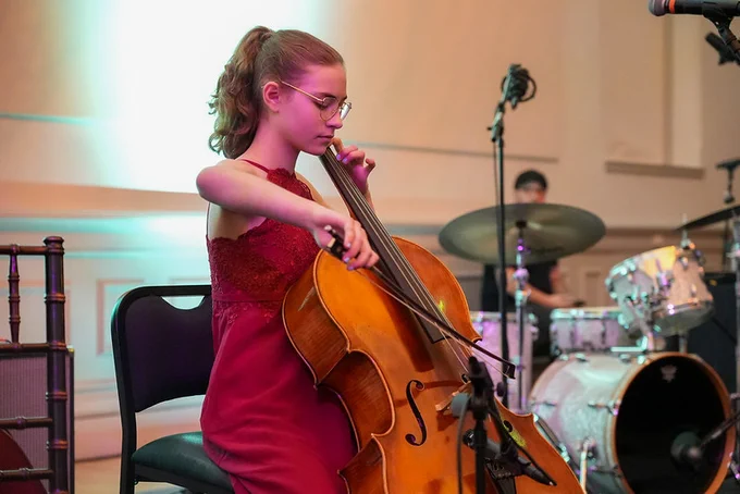 A student in a red dress sitting in an amphitheater, playing a strings instrument. Behind the student, a drum set is visible. 