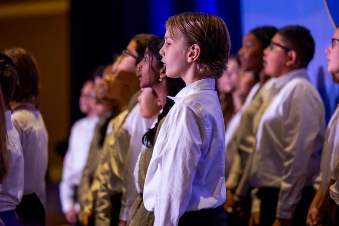 A group of young students in a choir are singing on stage while wearing white and gold shirts.