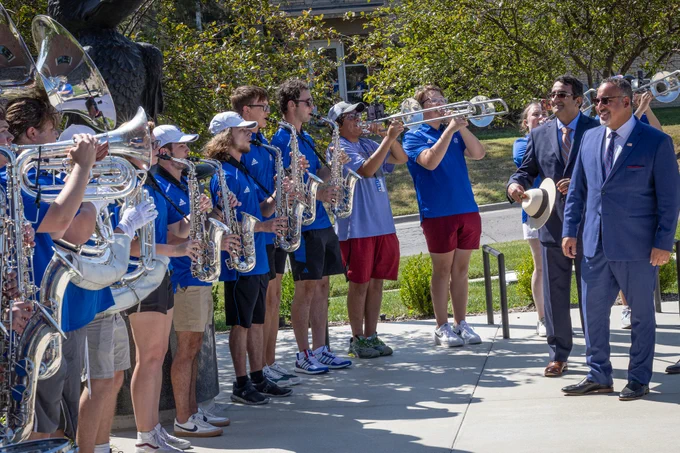 A group of students in blue shirts playing various band instruments in an outdoor campus space.