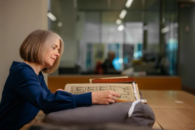 A photograph of a woman in a blue top looking at an old book filled with illustrations. The book is resting on a grey beanbag to protect it
