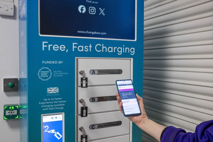 A photo of the new mobile phone charging unit. It's a rectangular box with a screen and has small cubby holes with keys for patients to lock their phone in securely. There is a person's hand and arm holding a phone in front of the unit. 