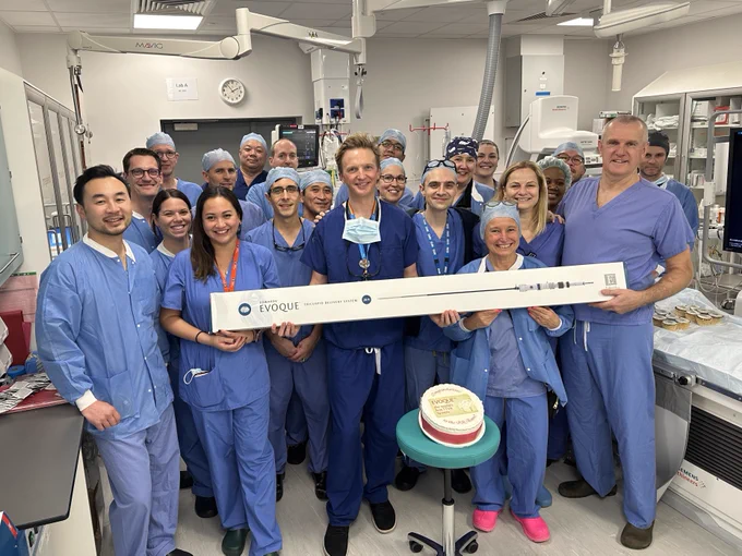 The team are pictured together smiling in the operating room with the equipment used in the procedure. There is a celebratory cake in front of them.