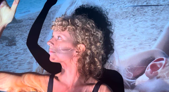 A girl with curly blond hair, in a blue tank top, has an image of the beach and her past self project on her face, body and the wall behind. 