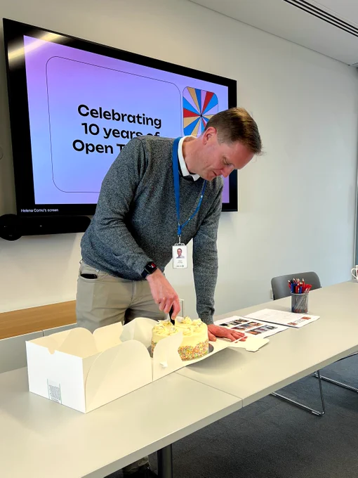 A man smiles as he cuts into a sprinkle-covered cake. Text on a screen behind him reads: celebrating 10 years of Open Targets.