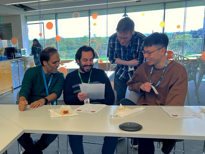 Four men laugh while looking at a sheet of paper. They are behind a desk in a meeting room. Green space with trees can be seen in the window behind them.