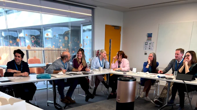 People sit in a meeting room, behind desks arranged at an angle. They are chatting with each other and laughing.