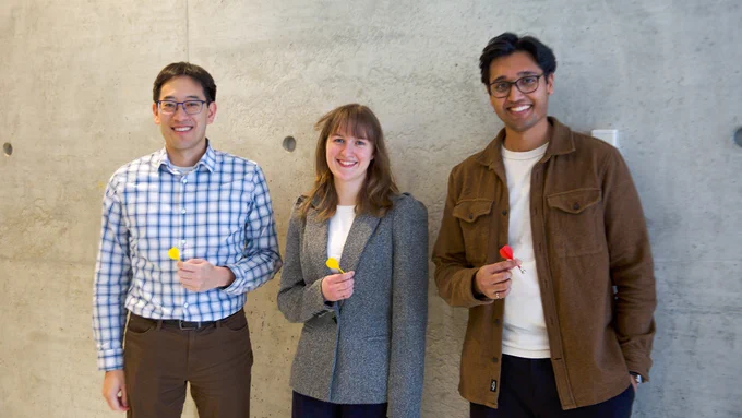 Three people stand in front of a concrete wall. Timothy Chan stands on the left wearing a shirt with a blue windowpane pattern and is holding a yellow dart, Rachael Walker stands in the centre wearing a grey blazer and is holding a yellow dart and Craig Fernandes stands on the right wearing a brown coat and is holding a red dart.