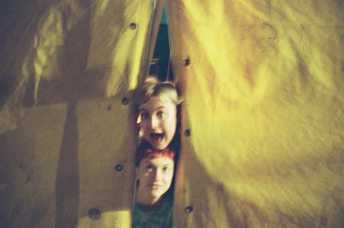 The entrance flaps to a yellow tent. Two smiling heads pop out of the entrance.