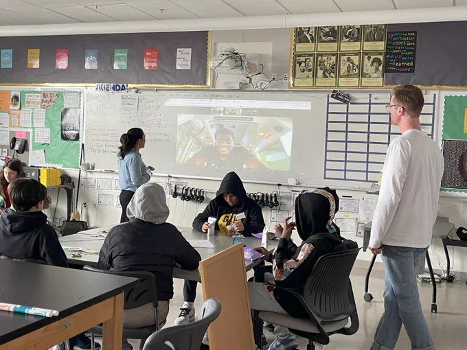 students sitting at desk in classrooms with adults standing and looking at projector screen