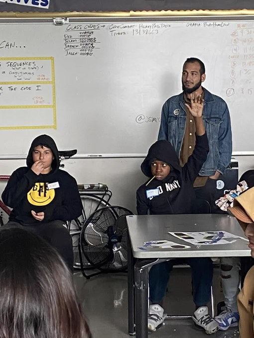 a student raising his hand at a desk, with another student sitting beside him and an adult standing behind them