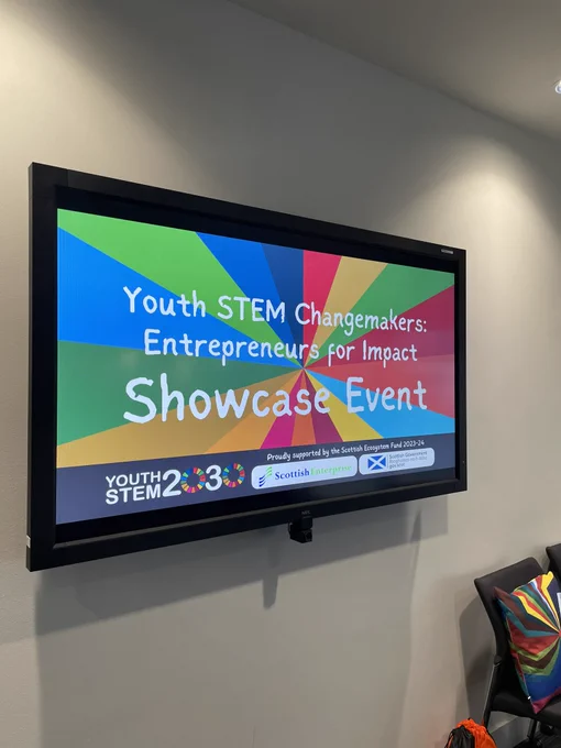 Screen on a wall with a slide reading ‘Youth STEM change makers entrepreneurs for impact showcase event’ The visuals are very colourful and multiple logos are visible along the bottom.