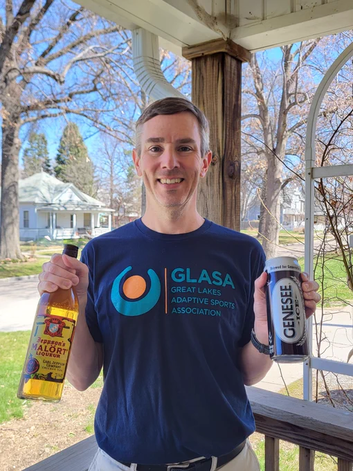 A man standing on a porch with a house in the background holds a bottle of Jeppson's Malort in his right hand and a 24-ounce can of Genesee ICE in his left hand. He is wearing a blue t-shirt that says, "Great Lakes Adaptive Sports Association."