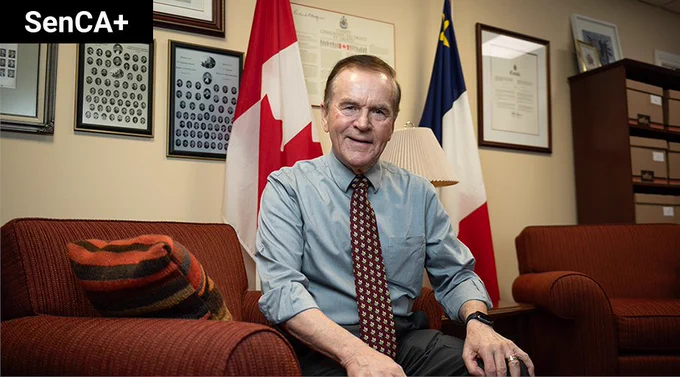 Senator Percy Mockler sits on a chair in his office with the Canadian and Acadian flags behind him.