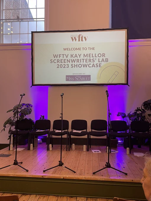 Stage set with three mics on stands. On the back wall, a screen reads, ‘Welcome to the WFTV Kay Mellor Screenwriters’ Lab 2023 Showcase.’