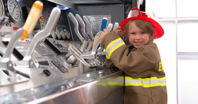 Child dressed up in a firefighter uniform playing in a decommissioned firetruck. 