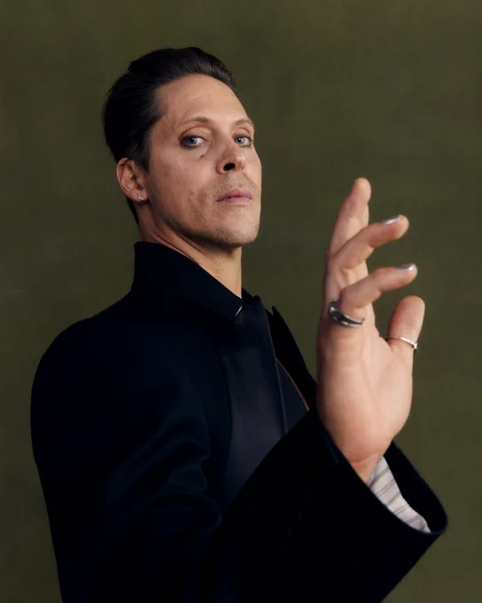 Neil Newbon poses in front of an earthy green backdrop in a striking black suit with his hand raised in front of the camera. He is looking down the lens seriously.