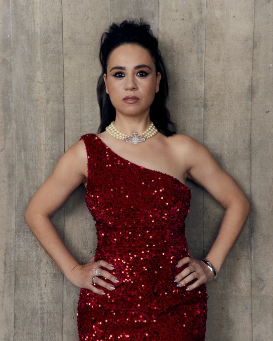 Samantha Beart poses in front of a wood panelled wall in a sparkly one shoulder red dress with both hands on their hips. They have a large pearl necklace on and their hair is in a half-up beehive-style do.