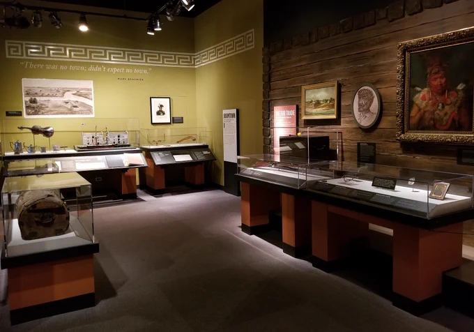 Color photograph of a museum gallery showing glass cases with objects & images hung on walls. 