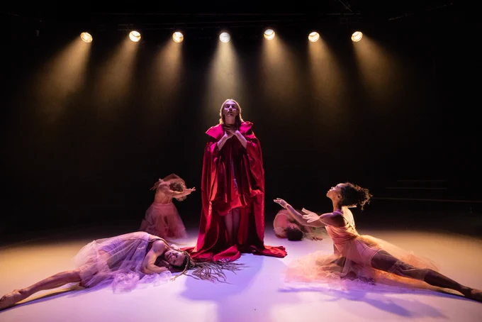 Jack Sears wearing a red cape surrounded by dancers lying on the floor wearing mesh dresses