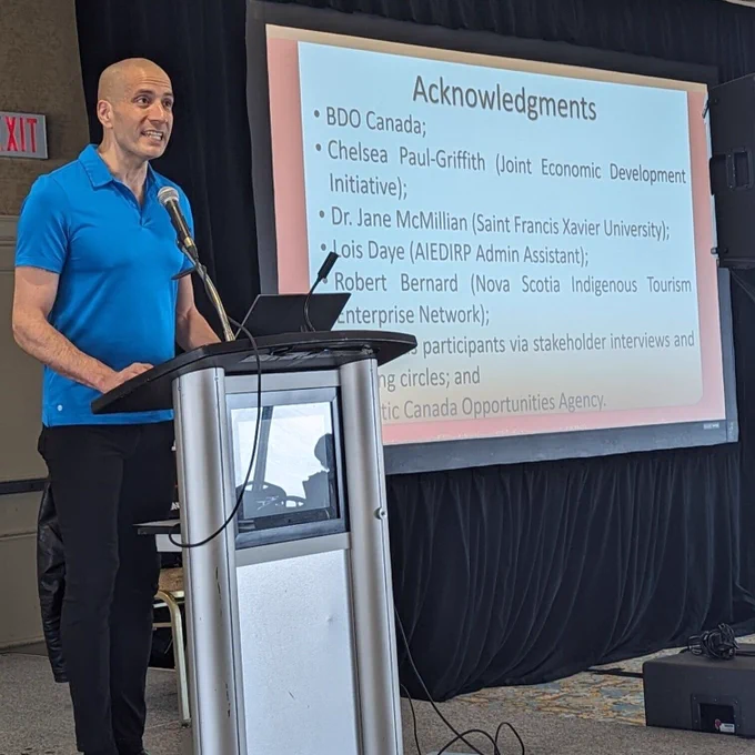 Jarvis Googoo presenting AIEDIRP Research program. He is standing in front of a podium on the left. On the right is a projector showing a slide.