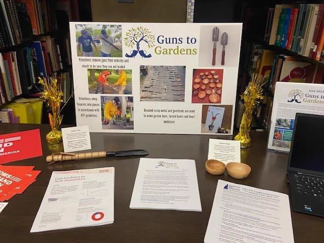 A photo of a table sign that says “Guns To Gardens” and has photos of guns being surrendered and turned into scrap metal and gardening tools. A small garden tool, Moms Demand red postcards, and pages of handouts that say “Gun Violence in New Hampshire” “Guns to Gardens” and “NH GVP Coalition Overview Session Year 2024” are placed on the table in front of the sign.