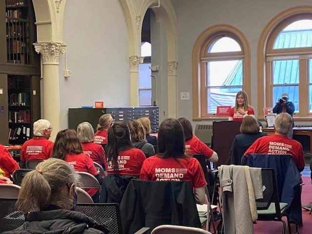 A photo of the backs of seated Moms Demand Action volunteers watching one person speak at a podium.