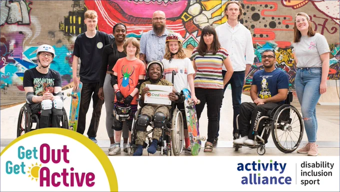 A group of disabled and none disabled people stand in a group posing for the camera. They are at a skate park, there's a backdrop of graffiti on a wall behind them. Some of the group are wearing helmets and knee and shin pads.