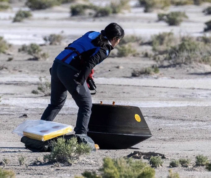 An engineer attends to the OSIRIS-REx sample return capsule soon after its landing in the Utah desert. The capsule is about the size of a large truck tire, charred black from reentry, and rests on flat, bare, salty soil with a few scrub plants nearby. 