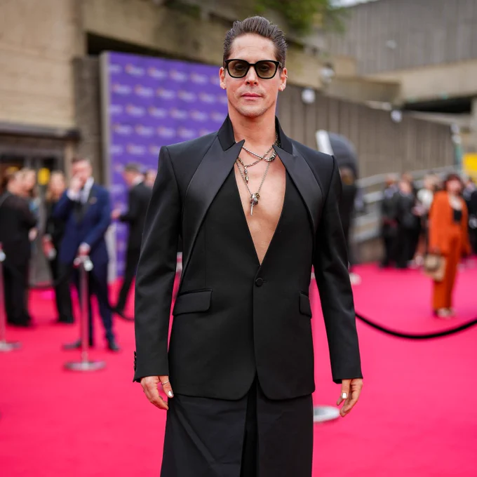 Neil Newbon attends the BAFTA Games Awards 2024 at the Queen Elizabeth Hall. He is in a striking-shaped v-neck black blazer, black trousers and sunglasses.