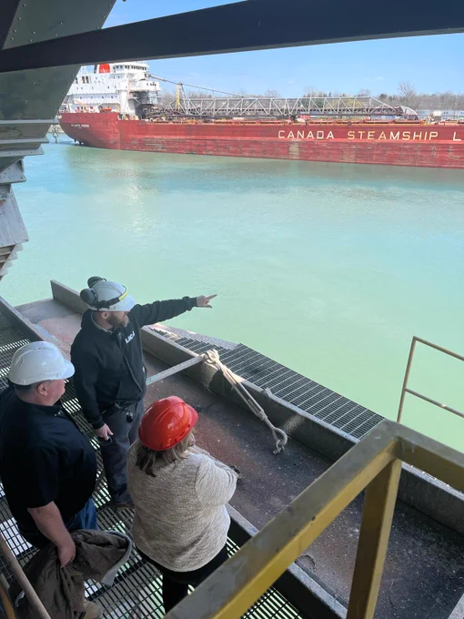 Two men and one female wearing hard hats look out over the port at a barge, while one points at something over the water.