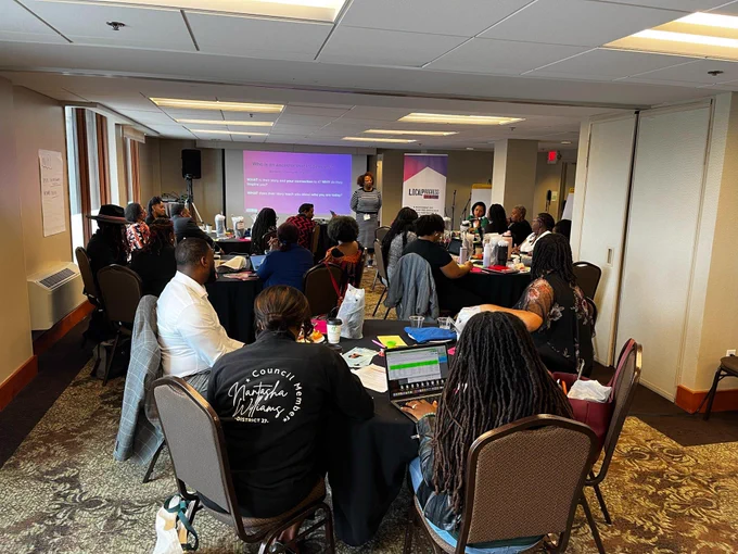 PHOTO: Group people in a meeting room. Signs in the background read "Local Progress Black Caucus"