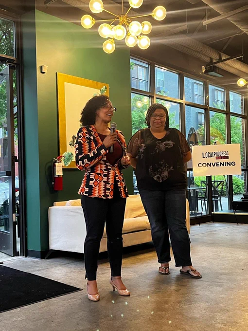 PHOTO: Two women standing next to each other. One is holding a microphone and address a crowd of people (not visible in picture).