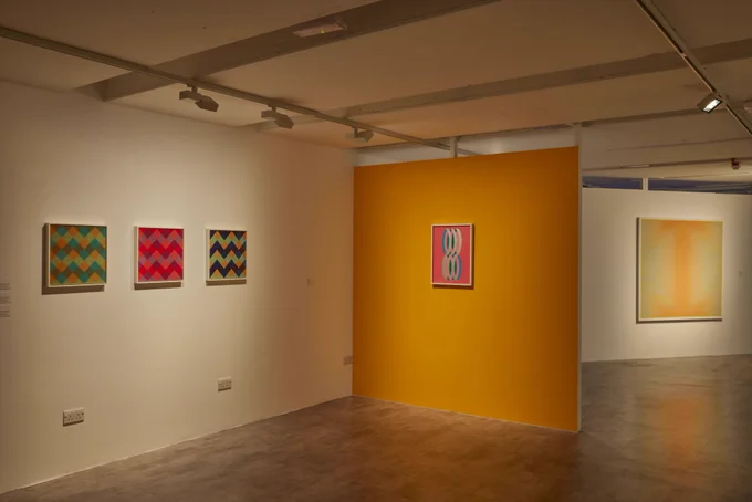 Moving Colour exhibition on display at The Winchester Gallery, WSA. Bright patterned artworks are displayed on white and a yellow ochre wall in a gallery space. Photo credit Dave Gibbons