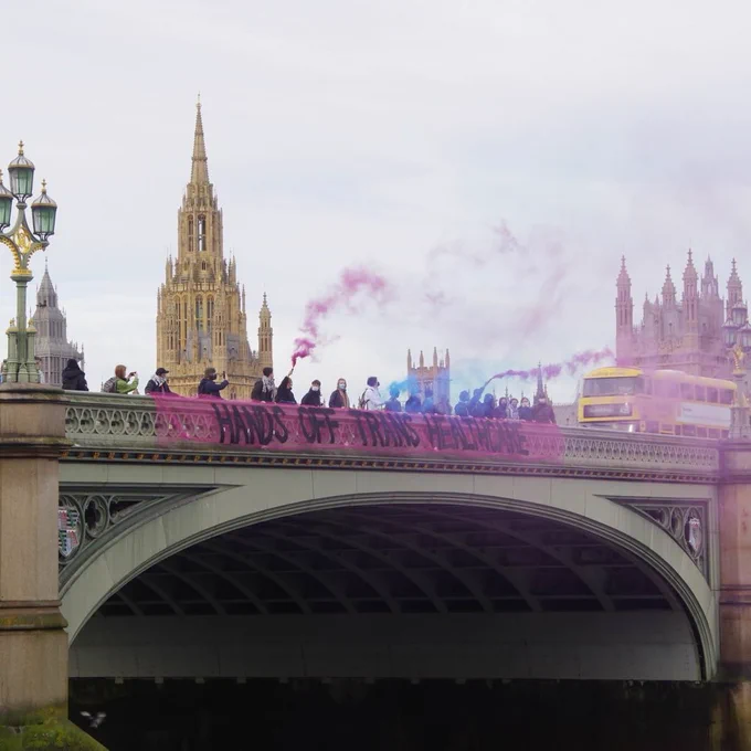 A group of 18 activists hang a banner reading 'Hands off trans healthcare' over the side of Westminster Bridge. The banner is pink mesh with black text. Pink, blue and purple smoke from flares fills the sky. In the background are the Houses of Parliament, and a double decker bus crossing the bridge.