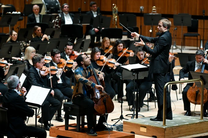 Cellist Sheku Kanneh-Mason in a gold and blue floral shirt performing on stage looking at Vasily conducting.