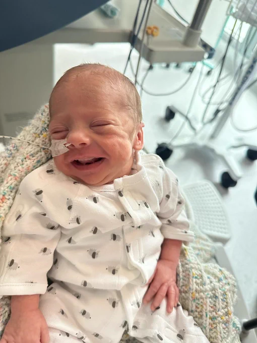 A baby is seen smiling while wearing the new sleep suit. They are in a hospital ward and have a small tube coming from their nose.