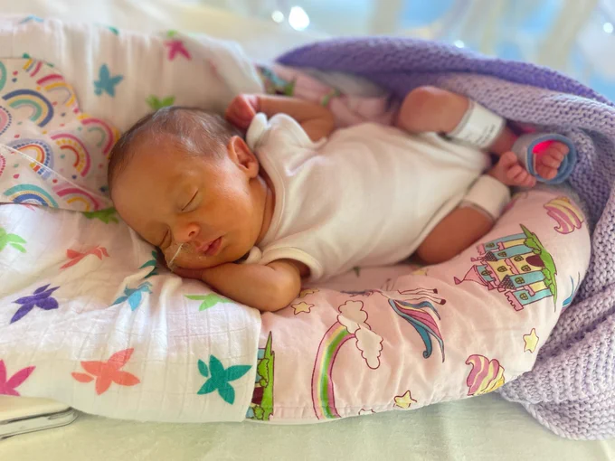 A baby is seen sleeping while wearing new baby clothes. They are in a small bed and have a small tube coming from their nose.