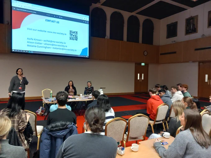 A presentation screen with slides, people seated on chairs and a person speaking