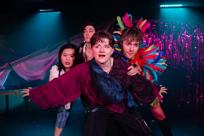 The cast of Pansexual Pregnant Piracy are clumped together with Ro Suppa at the front in an oversized maroon shirt, looking off invitingly to the side. Immediately over their shoulder Robbie Taylor Hunt is wearing colourful feathers on his head and across his shoulders. Behind the group are a pink shimmer curtain and pink fabric tied to a diagonal mast.